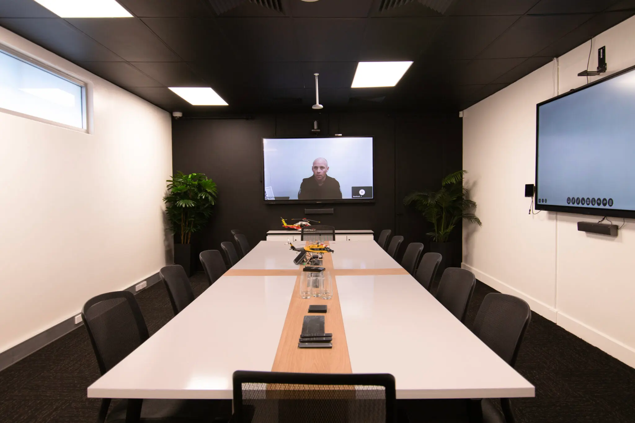 A white meeting table sits before a screen displaying a man in black, in a white room