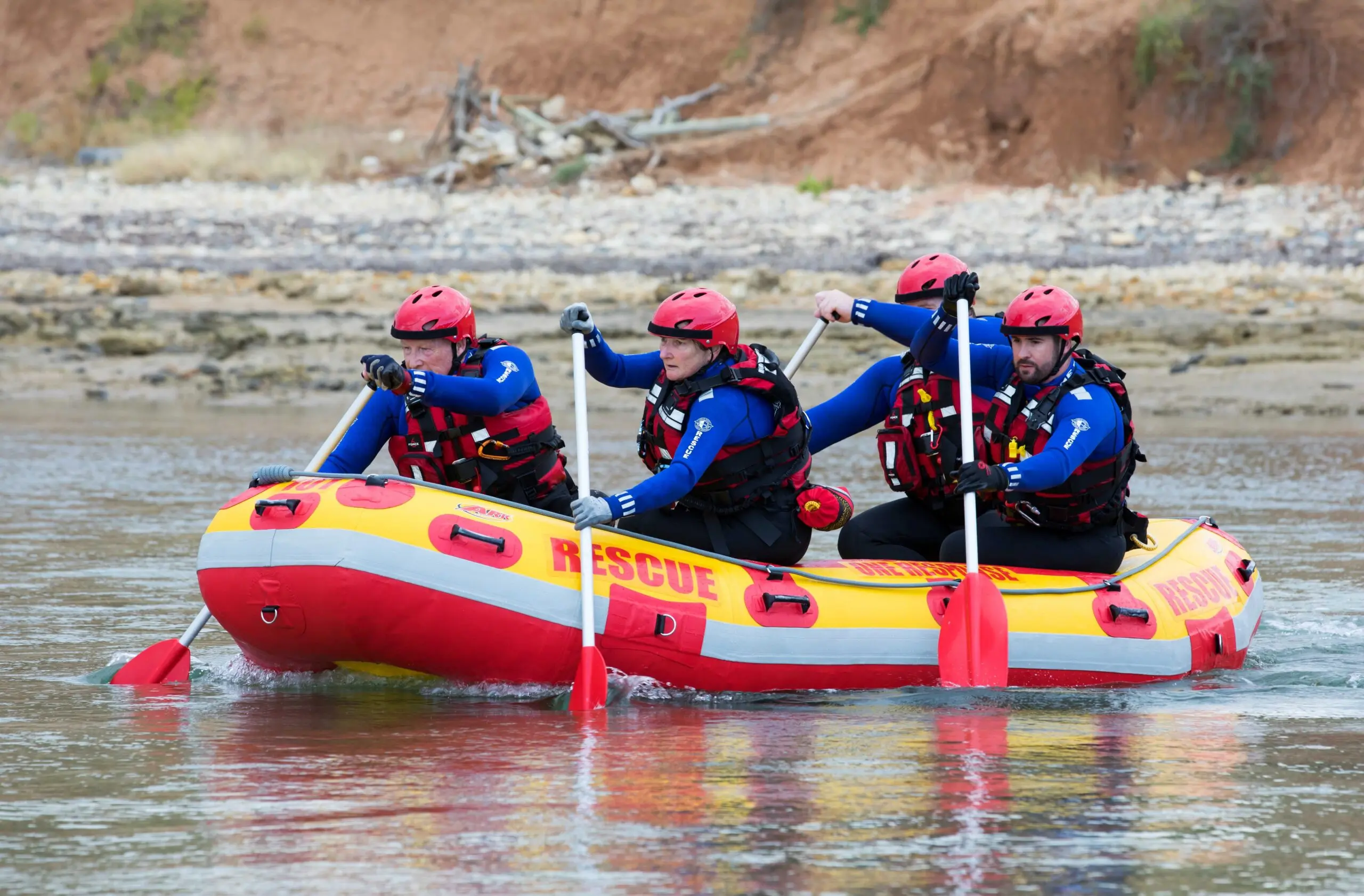 Four people — one wearing a blue shirt, one wearing a red vest, and one with a red hat — are paddling a red and yellow inflatable boat on a river.