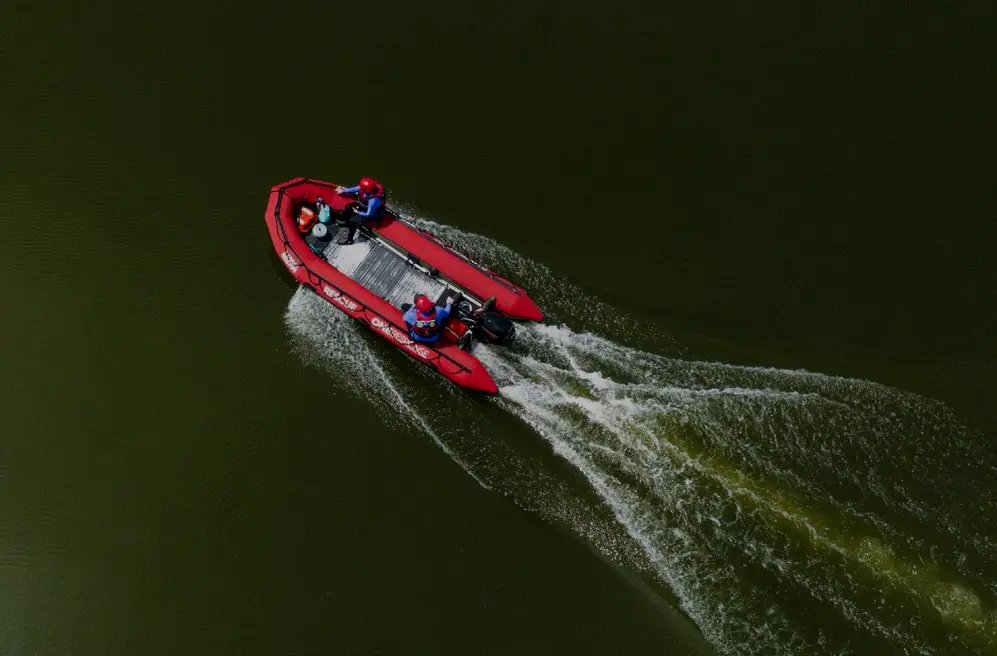 Two men in blue shirts, red vests, and red hats are sitting on a red inflatable boat in the middle of the water